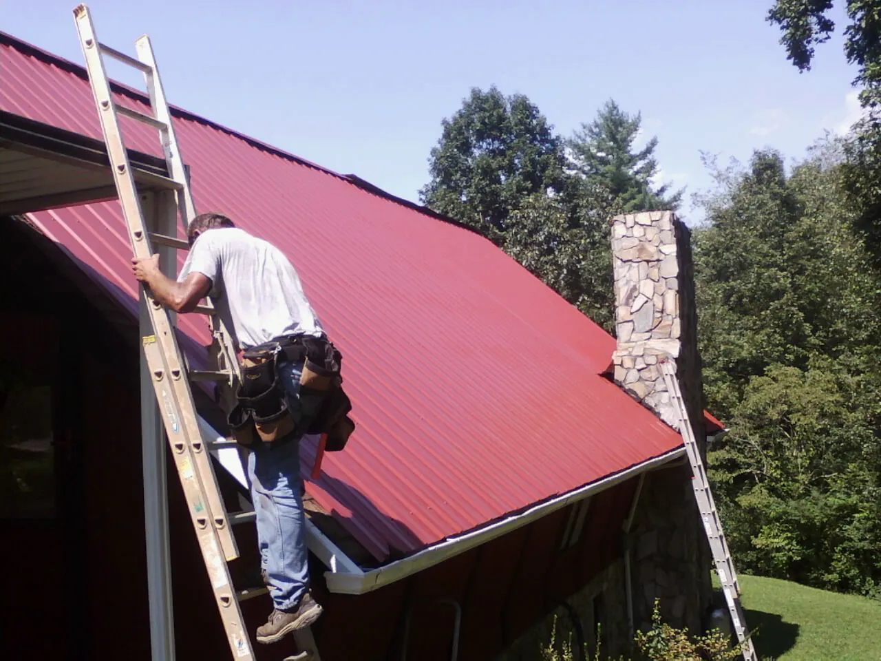 Roofer on a ladder beside a red metal roof and stone chimney — Soffit & Fascia Repair by Roofing Brothers in Faxon